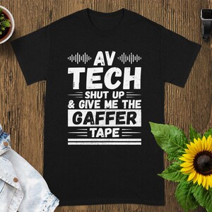 May include: Black t-shirt with white text that reads "AV TECH SHUT UP & GIVE ME THE GAFFER TAPE". The shirt is displayed on a wooden surface with a sunflower and a small potted plant.