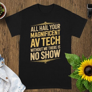 May include: Black t-shirt with the text "ALL HAIL YOUR MAGNIFICENT AV TECH WITHOUT ME THERE IS NO SHOW" in a gold color. The shirt is on a wooden surface with a small plant, a denim jacket, a sunflower, and a smartwatch.