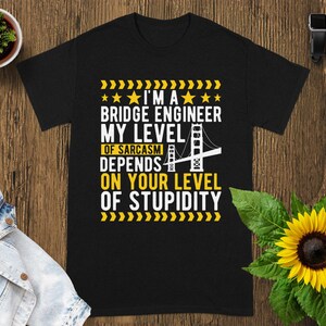 May include: A black short-sleeve t-shirt featuring yellow and white text that reads: I'M A BRIDGE ENGINEER MY LEVEL OF SARCASM DEPENDS ON YOUR LEVEL OF STUPIDITY. The design includes a graphic of a suspension bridge and yellow chevron patterns.