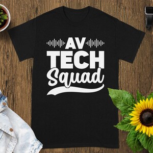 May include: Black t-shirt with the text "AV TECH Squad" in white, featuring a sound wave design above the words "AV" and below "Squad". The shirt is displayed on a wooden surface with a sunflower and a denim jacket.
