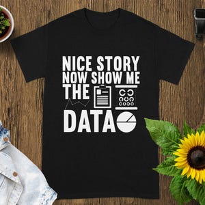 Peut inclure: T-shirt noir avec l'inscription blanche "NICE STORY NOW SHOW ME THE DATA". Le design comprend des icônes de document, de tableau ophtalmique et de camembert. Le t-shirt est posé sur une surface en bois, avec un tournesol et une petite plante.