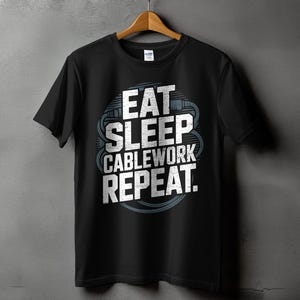 May include: Black t-shirt with the text "EAT SLEEP CABLEWORK REPEAT" in white, with a blue and gray cable design. The shirt is hanging on a wooden hanger against a gray wall.