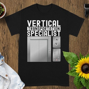 May include: Black t-shirt featuring the text "VERTICAL TRANSPORTATION SPECIALIST" above a silver elevator graphic with buttons. The shirt is displayed on a wooden surface.
