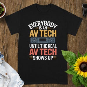 May include: Black t-shirt with the text "EVERYBODY IS AN AV TECH UNTIL THE REAL AV TECH SHOWS UP" in white, orange, and red. The design includes a graphic of a stereo. The shirt is displayed on a wooden surface.