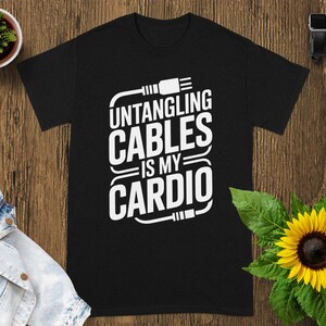 May include: Black t-shirt with the white text "UNTANGLING CABLES IS MY CARDIO". The shirt is displayed on a wooden surface, with a small potted plant, a denim jacket, a sunflower, and a smartwatch in the background.