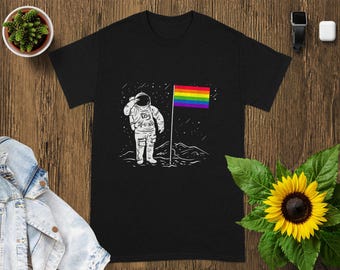 May include: Black t-shirt with a white illustration of an astronaut saluting a rainbow flag on a lunar surface. The design includes stars and a mountain range. The shirt is displayed on a wooden surface with a sunflower and a small plant.