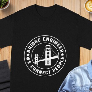 May include: A black short-sleeved t-shirt featuring a white circular graphic on the chest. The graphic depicts a suspension bridge with the text "BRIDGE ENGINEER" and "WE CONNECT PEOPLE" arranged in a circle around the bridge illustration.
