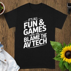 May include: Black t-shirt with the white text "IT'S ALL FUN & GAMES UNTIL YOU BLAME THE AV TECH". The shirt is on a wooden surface with a small potted plant, a denim jacket, a sunflower, a smartwatch, and a wireless earbud case.