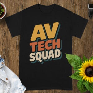 May include: Black t-shirt with the text "AV TECH SQUAD" in orange, red, and white, with a blue outline. The shirt is on a wooden surface, with a small potted plant, a denim jacket, a sunflower, a smartwatch, and a wireless earbud case.