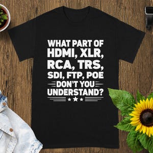 May include: Black t-shirt with white text: "WHAT PART OF HDMI, XLR, RCA, TRS, SDI, FTP, POE DON'T YOU UNDERSTAND?" The shirt is on a wooden surface with a sunflower and a small potted plant.