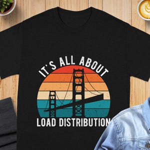 May include: A black short-sleeve t-shirt featuring a graphic of the Golden Gate Bridge silhouette against a retro-style sunset of orange, yellow, and teal stripes. The text "IT'S ALL ABOUT LOAD DISTRIBUTION" is printed in white capital letters.