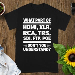 May include: Black t-shirt with white text: "WHAT PART OF HDMI, XLR, RCA, TRS, SDI, FTP, POE DON'T YOU UNDERSTAND?" The shirt is on a wooden surface, with a small potted plant, a denim jacket, a sunflower, and a smartwatch.