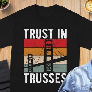 May include: A black short-sleeve t-shirt featuring a graphic of the Golden Gate Bridge silhouette over horizontal stripes in red, orange, yellow, and light blue. The text "TRUST IN TRUSSES" is printed in white capital letters above and below the graphic.