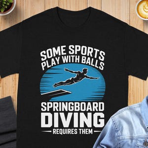 May include: Black t-shirt with a graphic of a diver in mid-air above a diving board, set against a blue and white background. Text reads "SOME SPORTS PLAY WITH BALLS SPRINGBOARD DIVING REQUIRES THEM".