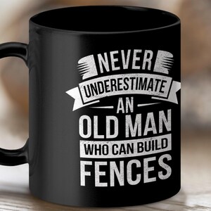 Op de afbeelding: Een zwarte keramische mok met witte tekst die luidt: NEVER UNDERESTIMATE AN OLD MAN WHO CAN BUILD FENCES. Het ontwerp bevat een grafische banner boven het woord UNDERESTIMATE en gestileerde letters voor de overige woorden.
