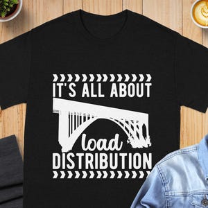 May include: A black short-sleeve t-shirt featuring a white graphic of an arched bridge. The text above the bridge reads "IT'S ALL ABOUT" and the text below reads "load DISTRIBUTION" in a mix of block and script fonts, framed by chevron patterns.