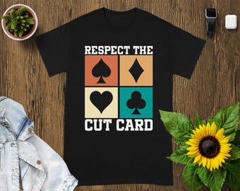 May include: Black t-shirt with the text "RESPECT THE CUT CARD" above four playing card suit symbols: spade, diamond, heart, and club. The shirt is on a wooden surface with a small plant and a sunflower.