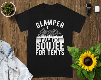 Peut inclure: T-shirt noir avec texte blanc "GLAMPER WAY TOO BOOJEE FOR TENTS". Le motif comprend une chaîne de montagnes, un croissant de lune et des lignes horizontales. Un tournesol et une petite plante en pot sont en arrière-plan.