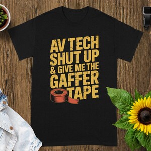 May include: Black t-shirt with the text "AV TECH SHUT UP & GIVE ME THE GAFFER TAPE" in yellow. A roll of red gaffer tape is featured below the text. The shirt is displayed on a wooden surface with a small potted plant and a sunflower.