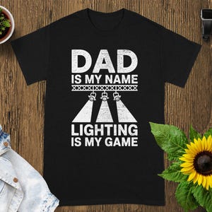 Puede incluir: Camiseta negra con el texto blanco "DAD IS MY NAME LIGHTING IS MY GAME". El diseño incluye focos de escenario. La camiseta se muestra sobre una superficie de madera con una pequeña planta en maceta, una chaqueta vaquera, un girasol y un reloj inteligente.
