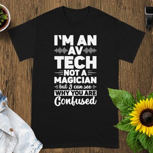 May include: Black t-shirt with the text "I'M AN AV TECH NOT A MAGICIAN but I can see WHY YOU ARE Confused" in white. The shirt is on a wooden surface with a sunflower and a small potted plant.