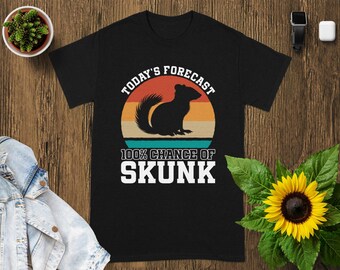 May include: Black t-shirt with a graphic of a skunk inside a sunset circle. The text reads "Today's Forecast" above the skunk and "100% Chance of Skunk" below. The shirt is on a wooden surface.