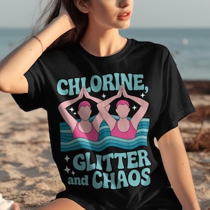 Puede incluir: Camiseta negra con el texto "CHLORINE, GLITTER and CHAOS" en azul claro y blanco. El gráfico presenta dos figuras con trajes de baño rosas en el agua, con las manos levantadas. La camiseta la lleva una persona.