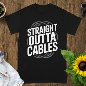 May include: Black t-shirt with the text "STRAIGHT OUTTA CABLES" in white, encircled by a gray cable design. The shirt is displayed on a wooden surface, with a small potted plant, a denim jacket, a sunflower, and a smartwatch.