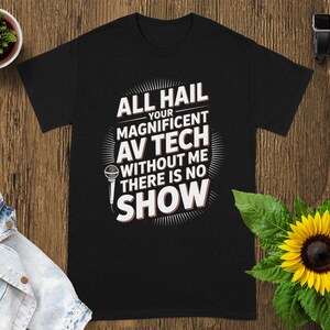 May include: Black t-shirt with the text "ALL HAIL YOUR MAGNIFICENT AV TECH WITHOUT ME THERE IS NO SHOW" in white. The design includes a microphone graphic. The shirt is displayed on a wooden surface with a sunflower and a small plant.