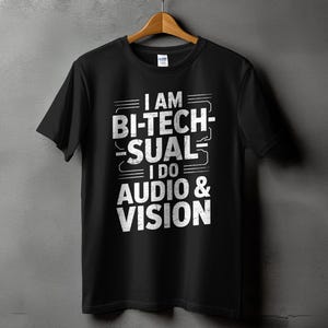May include: Black t-shirt with the text "I AM BI-TECH-SUAL I DO AUDIO & VISION" in white, hanging on a wooden hanger against a gray wall. The shirt is a Gildan brand.