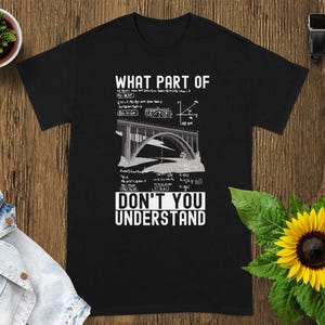 May include: Black t-shirt with the text "WHAT PART OF DON'T YOU UNDERSTAND" in white. The design includes a bridge illustration and mathematical equations. The shirt is laid on a wooden surface.