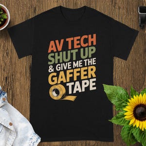 May include: Black t-shirt with the text "AV TECH SHUT UP & GIVE ME THE GAFFER TAPE" in orange, green, yellow, and white. A roll of tape is illustrated below the text. The shirt is displayed on a wooden surface.