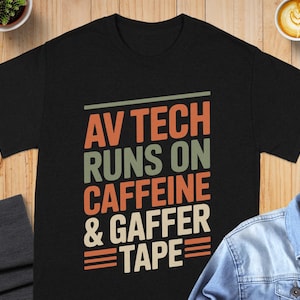 May include: Black t-shirt with the text "AV TECH RUNS ON CAFFEINE & GAFFER TAPE" in orange, green, and white. The shirt is displayed on a wooden surface with a denim jacket, a cup of coffee, and a small plant.