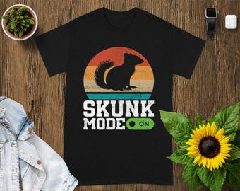 May include: Black t-shirt with a retro design featuring a skunk silhouette inside a sunset circle. The text "SKUNK MODE ON" is printed below the graphic in white. The design has a vintage, outdoorsy feel.