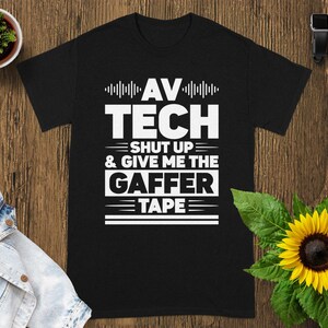 May include: Black t-shirt with white text that reads "AV TECH SHUT UP & GIVE ME THE GAFFER TAPE". The shirt is displayed on a wooden surface with a sunflower and a small potted plant.