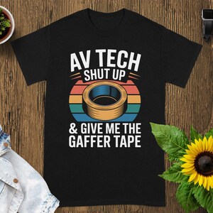 May include: Black t-shirt with the text "AV TECH SHUT UP & GIVE ME THE GAFFER TAPE". The design includes a roll of tape in front of a sunset-colored circle. A small potted plant and a sunflower are in the background.