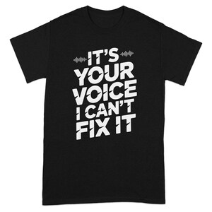May include: Black t-shirt with the white text "IT'S YOUR VOICE I CAN'T FIX IT". The text is arranged in a stacked format, with a sound wave graphic above the first line. The shirt is made of a soft material.