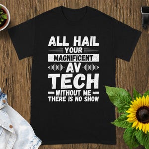 May include: Black t-shirt with white text that reads "ALL HAIL YOUR MAGNIFICENT AV TECH WITHOUT ME THERE IS NO SHOW." The shirt is on a wooden surface with a sunflower and a small potted plant.