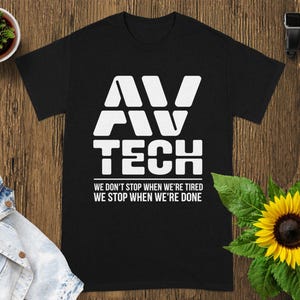May include: Black t-shirt with the white text "AV TECH" above the quote "WE DON'T STOP WHEN WE'RE TIRED WE STOP WHEN WE'RE DONE." The shirt is displayed on a wooden surface with a sunflower and a small potted plant.