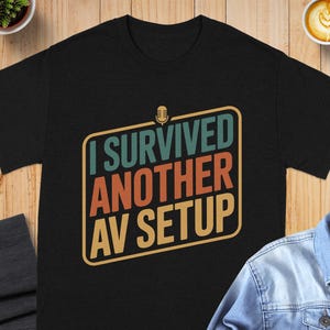 May include: Black t-shirt with the text "I Survived Another AV Setup" in a retro-style design. The text is in teal, orange, and yellow, with a microphone graphic. The shirt is on a wooden surface with a coffee cup and a small plant.