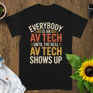 May include: Black t-shirt with the text "EVERYBODY IS AN AV TECH UNTIL THE REAL AV TECH SHOWS UP" in a distressed, vintage style. The text is in white, orange, and yellow. The shirt is on a wooden surface.