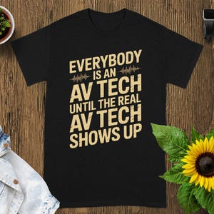 May include: Black t-shirt with the text "EVERYBODY IS AN AV TECH UNTIL THE REAL AV TECH SHOWS UP" in a yellow font. The shirt is displayed on a wooden surface with a sunflower and a small potted plant.
