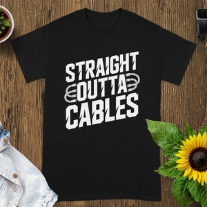 May include: Black t-shirt with the white text "STRAIGHT OUTTA CABLES". The shirt is displayed on a wooden surface, with a small potted plant, a denim jacket, a sunflower, a smartwatch, and wireless earbuds.