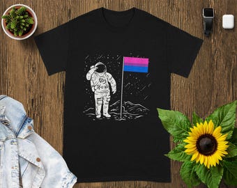 May include: Black t-shirt with a white illustration of an astronaut saluting a flag with pink, purple, and blue stripes. The design is set against a starry sky and a lunar landscape.