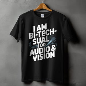 May include: Black t-shirt with the text "I AM BI-TECHSUAL I DO AUDIO & VISION" in white, with a microphone graphic. The shirt is hanging on a wooden hanger against a gray wall.