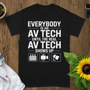 May include: Black t-shirt with the text "EVERYBODY is an AV TECH until the REAL AV TECH SHOWS UP" in white. The shirt has three small icons below the text. The shirt is on a wooden surface.