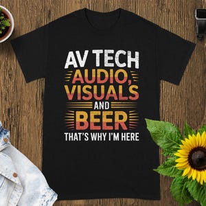May include: Black t-shirt with the text "AV TECH AUDIO, VISUALS AND BEER THAT'S WHY I'M HERE" in white, orange, and yellow. The shirt is displayed on a wooden surface with a small potted plant, a denim jacket, a sunflower, and a smartwatch.
