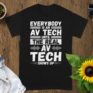 May include: Black t-shirt with the white text "EVERYBODY IS AN AV TECH UNTIL THE REAL AV TECH SHOWS UP." The shirt is displayed on a wooden surface with a small potted plant, a denim jacket, a sunflower, a smartwatch, and a white square.