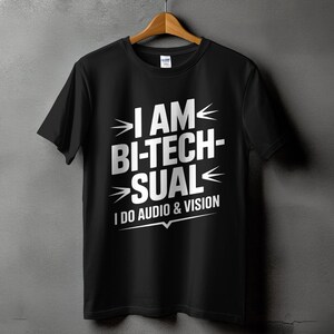 May include: Black t-shirt with the text "I AM BI-TECHSUAL I DO AUDIO & VISION" in white. The shirt is hanging on a wooden hanger against a gray wall. The text is in a bold, modern font.