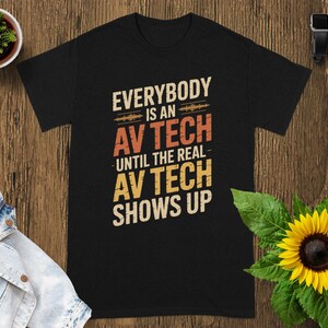 May include: Black t-shirt with the text "EVERYBODY IS AN AV TECH UNTIL THE REAL AV TECH SHOWS UP" in white, orange, and yellow. The shirt is on a wooden surface with a sunflower and a small potted plant.